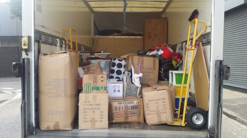 Inside a professionally maintained vehicle used by Commercial Waste Addiscombe, there is an organized assortment of various types of domestic and light commercial rubbish. The cargo area is filled with large cardboard boxes, some securely taped and others open, containing an array of waste materials such as paper waste, packaging, and household items, with visible print and labels on several boxes. Leaning against the sides of the vehicle are two yellow hand trucks or dollies, one on each side, equipped with black rubber wheels and handles, used for manual transport of heavy or bulky items. The background reveals a partially visible interior wall of the vehicle, finished with a smooth, light-colored interior lining. The scene appears to be set on a typical urban street or driveway, with a glimpse of a closed grey metal roller shutter on the right side, suggesting a commercial area or service yard in the vicinity of Addiscombe, within the postcode region associated with south London. The lighting is diffuse, indicating an overcast day, and the overall environment emphasizes the logistics and waste management task performed by Commercial Waste Addiscombe in handling rubbish collection efficiently and systematically.