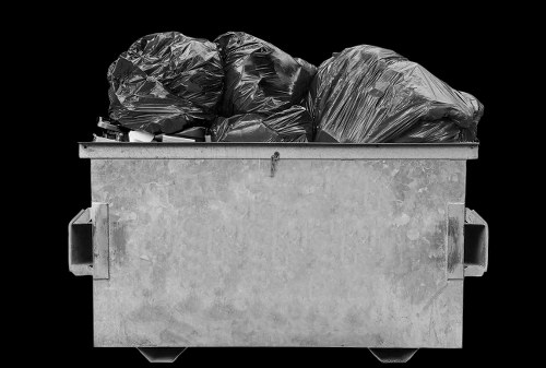 A large, rectangular, grey plastic waste skip situated outdoors on a paved surface, filled with multiple black heavy-duty rubbish bags made of thick plastic, some with a crinkled or tied top, stacked and slightly leaning against each other. The skip features two built-in handles on each side and rests against a solid black background, emphasizing its utilitarian design. The environmental setting suggests a commercial or industrial area, possibly in Addiscombe or nearby London suburbs, consistent with local rubbish removal services such as Commercial Waste Addiscombe. The image captures the practical aspect of waste collection for business waste management, with clear details of the materials and textures of the bags and the sturdy construction of the skip itself, further aligning with rubbish disposal and collection activities typical for the postcode region.