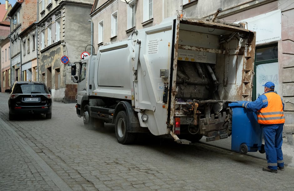 A sanitation worker wearing a yellow and red uniform is standing beside the rear of a large red waste collection vehicle on a street, actively loading rubbish into the open compartment. The vehicle's 