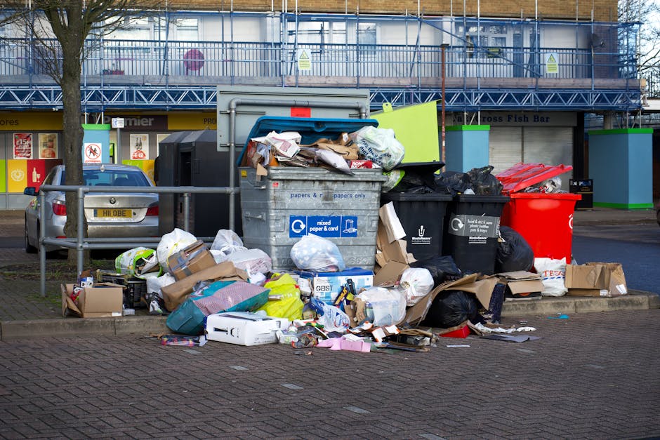 A street scene featuring a white waste collection truck actively engaged in rubbish removal on a cobblestone-paved road adjacent to older, multi-story buildings with mixed textures of brick and plaste