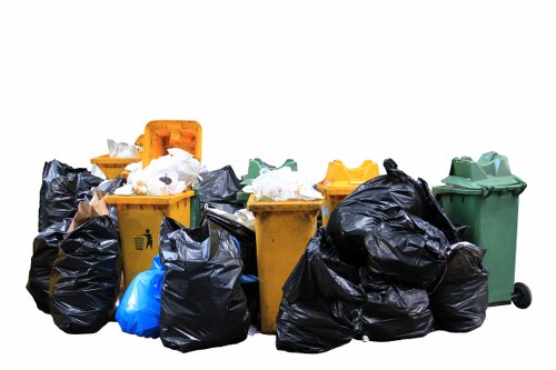 A collection of multiple wheelie bins and large black refuse bags placed outdoors on a paved surface, likely part of a commercial waste collection scene; the bins include a yellow bin with an open lid containing white and black rubbish, a green bin to the far right with the lid closed, and other bins in yellow and black, some of which are partially obscured or leaning. The black rubbish bags are piled around and on top of the bins, appearing full and loosely tied, with some bags showing creases and glossy textures. A blue bag is visible among the black bags. The background is plain white, emphasizing the waste containers and bags, which are positioned in a compact arrangement, typical of a rubbish removal or waste disposal setting in an urban or suburban environment consistent with the area served by Commercial Waste Addiscombe, close to Croydon within the postcode area inferred from Addiscombe details.