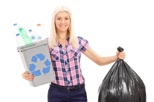 A young woman with blonde hair and a cheerful expression is standing against a plain white background, holding a small grey recycling bin filled with various plastic bottles and containers in her left hand. The bin features a prominent blue recycling symbol on the front. Her right hand is grasping a large black rubbish bag that appears to be full and tightly sealed at the top. She is dressed in a casual, short-sleeved, pink and blue checkered shirt and dark trousers. The scene emphasizes waste collection and rubbish disposal, aligning with the services provided by Commercial Waste Addiscombe. The background is neutral, keeping the focus on the woman and the waste items she is holding, which visually represent household or commercial waste suitable for professional rubbish removal in the Addiscombe area, within the postal code region serviced by the company.