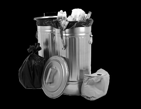 A stainless steel rubbish bin with a partially open lid, filled with crumpled paper, plastic bottles, and other waste materials, sits on a dark surface. To the left of the bin, there is a tied black plastic rubbish bag, and leaning against the bin is its detached lid. On the right side, a crumpled paper bag rests on the surface. The background appears plain and dark, emphasizing the metallic finish of the bin and the waste items. This setup reflects typical waste disposal and rubbish collection scenarios, consistent with professional rubbish removal services that might operate in Addiscombe or nearby areas. The image conveys a professional and straightforward approach to waste management, aligning with services offered by Commercial Waste Addiscombe for commercial or domestic waste clearance. The lighting highlights the reflective surface of the metal bin and the textures of the waste items, creating a clear, detailed visual of common rubbish receptacles and associated trash elements, suitable for illustrating waste management topics in the local context.